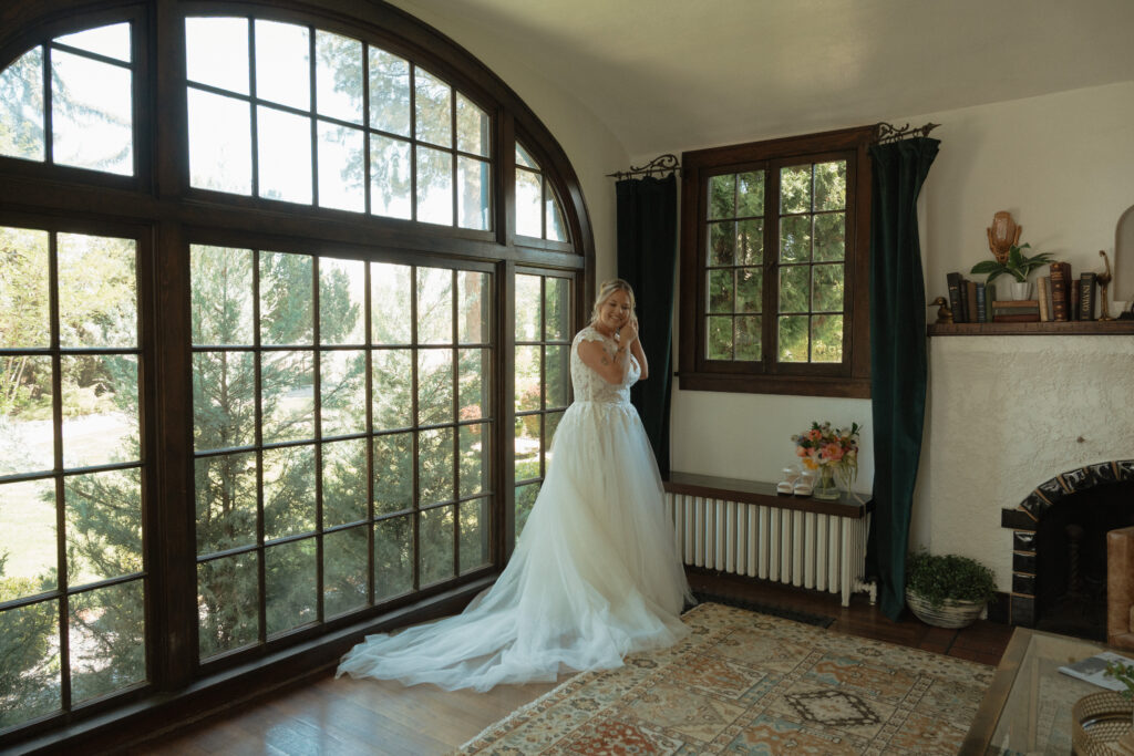 Bride in lace wedding gown adjusting earrings by window at The Cottage at River Bend in Eagle, Idaho.